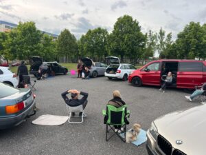 mehrer Menschen sitzen neben oder vor ihren autos, welche im Kreis stehen und die hunde sind im Auto oder neben den hundehaltern und schauen zu