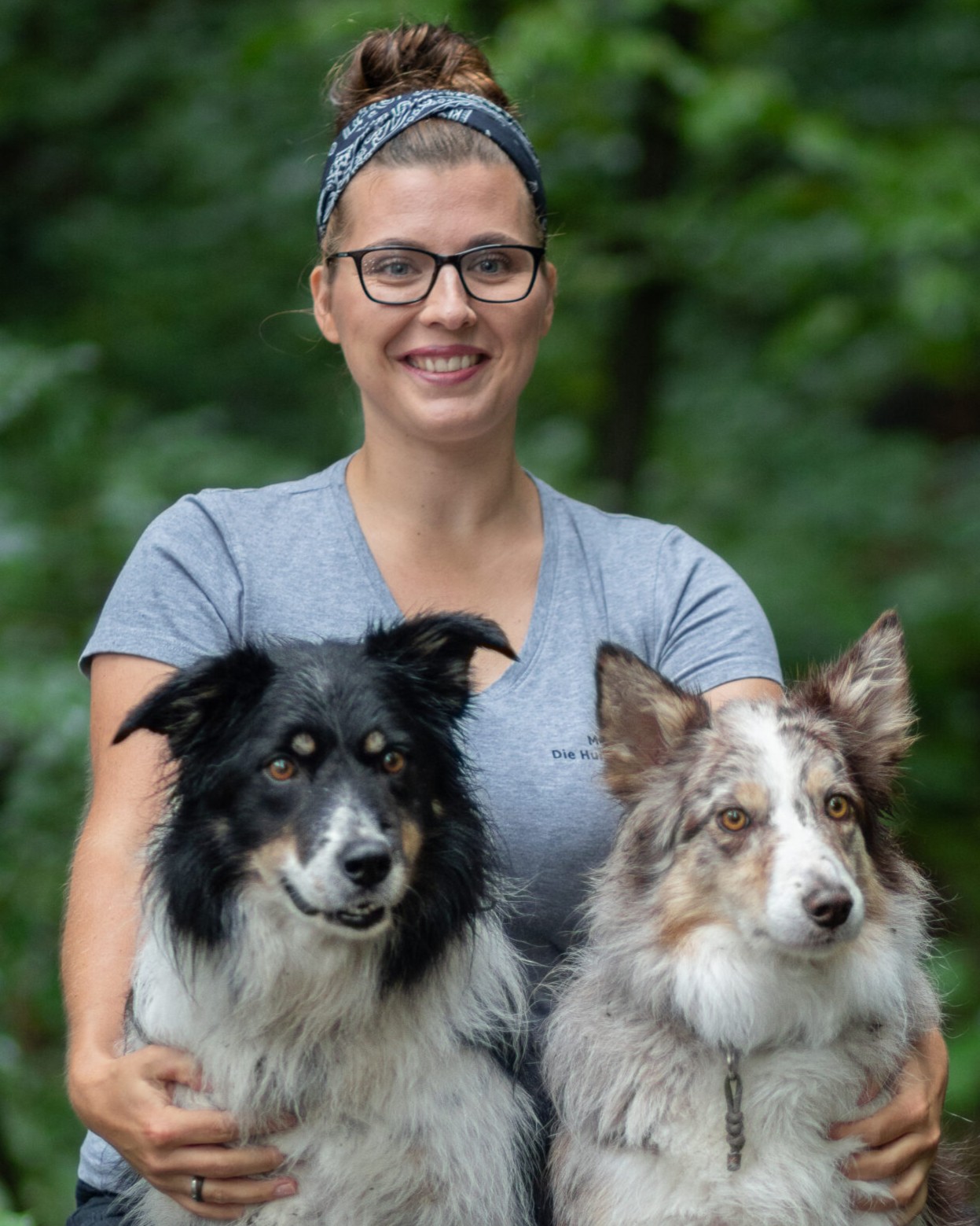 Hundetrainerin Maria Kurz lächelt mit ihren Hündinnen Cathy und Dörte vor grünem Laub