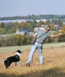 Hundelehrer Torsten kurz steht auf einem Feld und zeigt seitlich nach rechts gebeugt in eine Richtung. die hündin schaut ihn an und wartet auf ein zeichen zum start