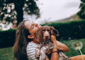 eine frau sitzt auf einer wiese, hat den ihren hund auf dem arm, hat die augengeschlossn und wünscht sich etwas. gleichzeitig weht der wind den samen einer pusteblume davon. sie wünscht sich etwas